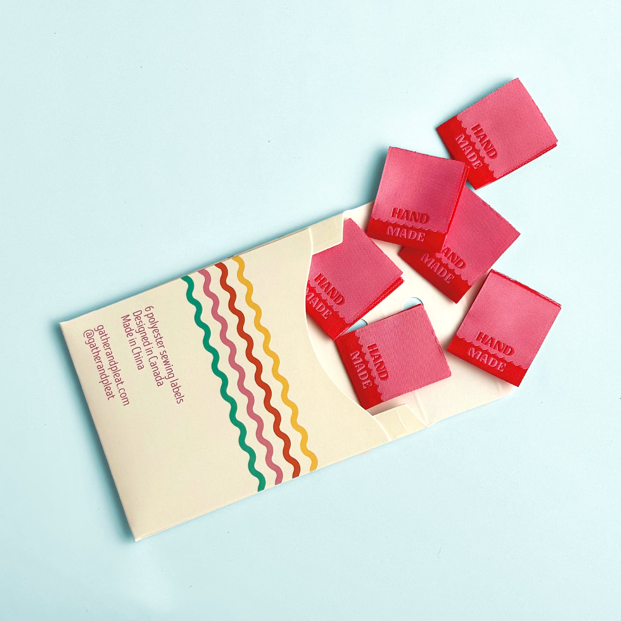 Pink and red 'Hand Made' woven sewing labels displayed beside opened Gather & Pleat packaging showing all six labels included in the pack.