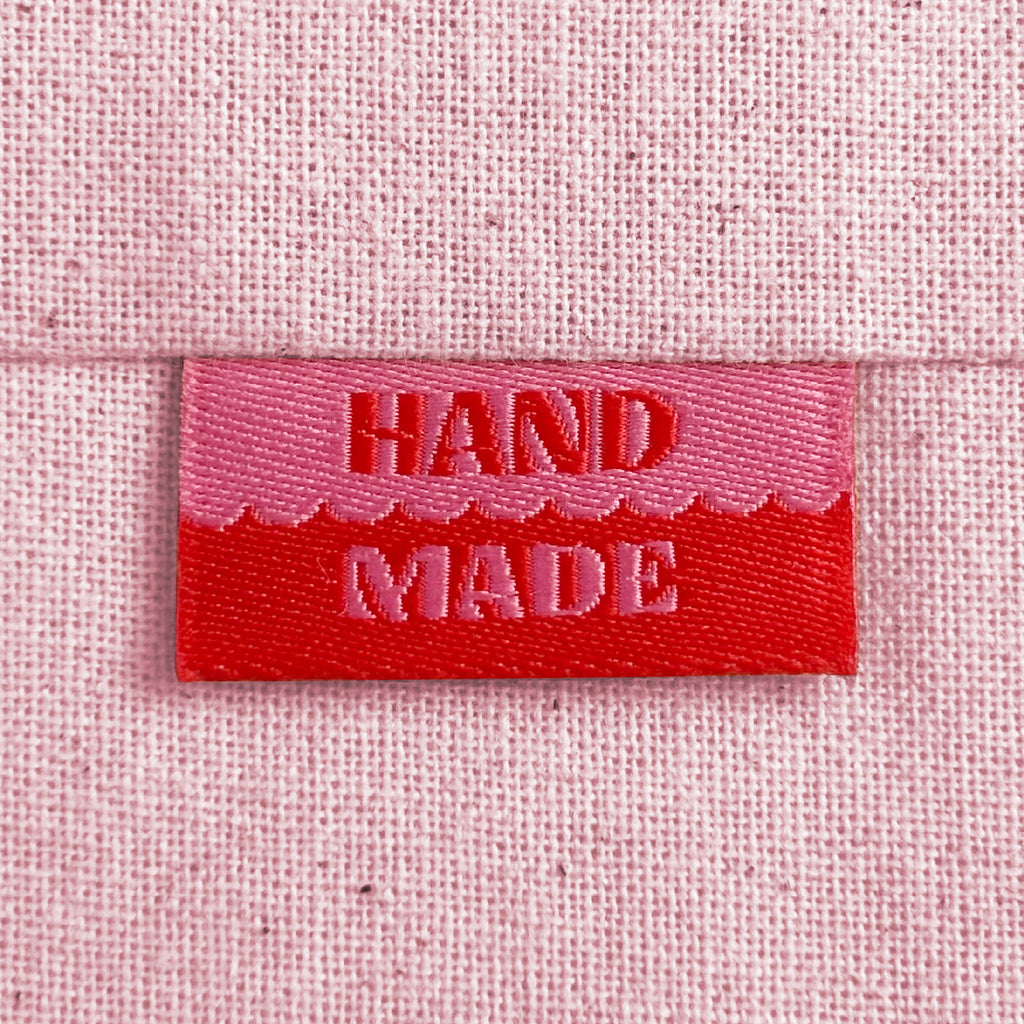A close-up of a pink and red woven sewing label with the words 'HAND MADE', sewn into light pink canvas.