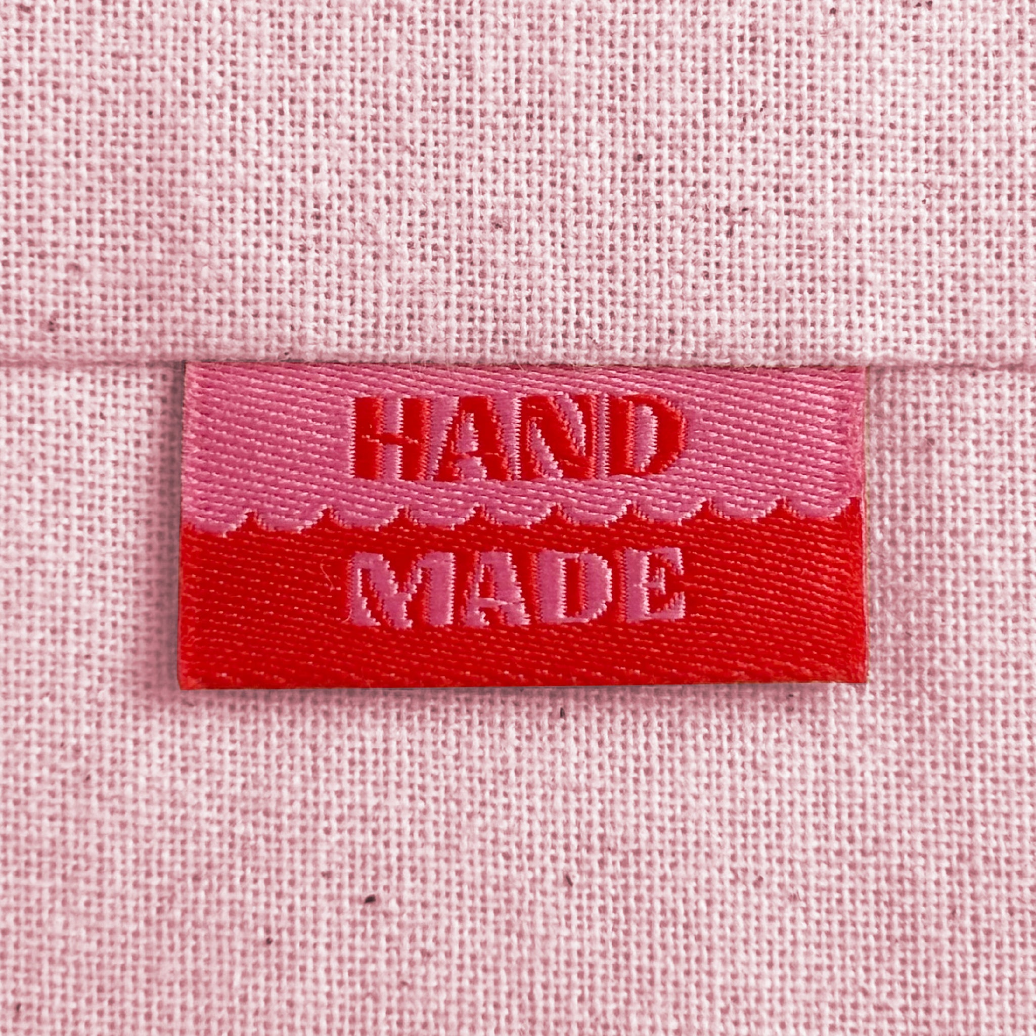 A close-up of a pink and red woven sewing label with the words 'HAND MADE', sewn into light pink canvas.