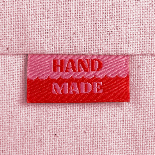 A close-up of a pink and red woven sewing label with the words 'HAND MADE', sewn into light pink canvas.