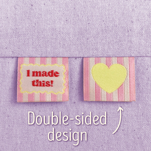 Two pink-striped, woven sewing labels that have been sewn into lilac canvas. The left label reads “I made this!” in red lettering, surrounded by a scalloped yellow border. The right label shows a yellow heart on a pink-striped background. Text in the image explains that this is a double-sided design, implying that the heart is on the back of the 'I Made This!' design.

