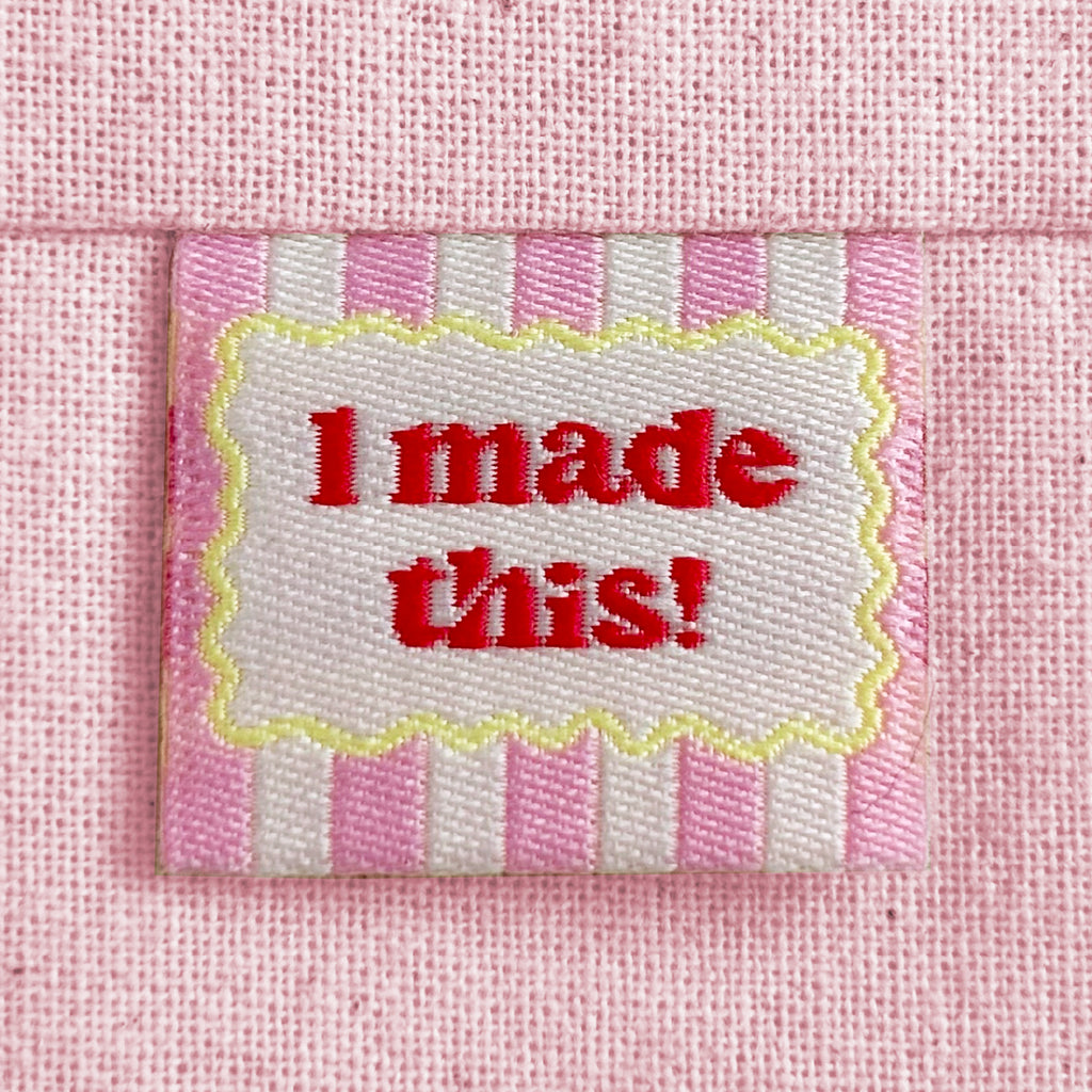 A close-up of a pink striped woven sewing label with a scalloped yellow border around red text reading 'I made this!'. The label is sewn into a light pink canvas fabric.