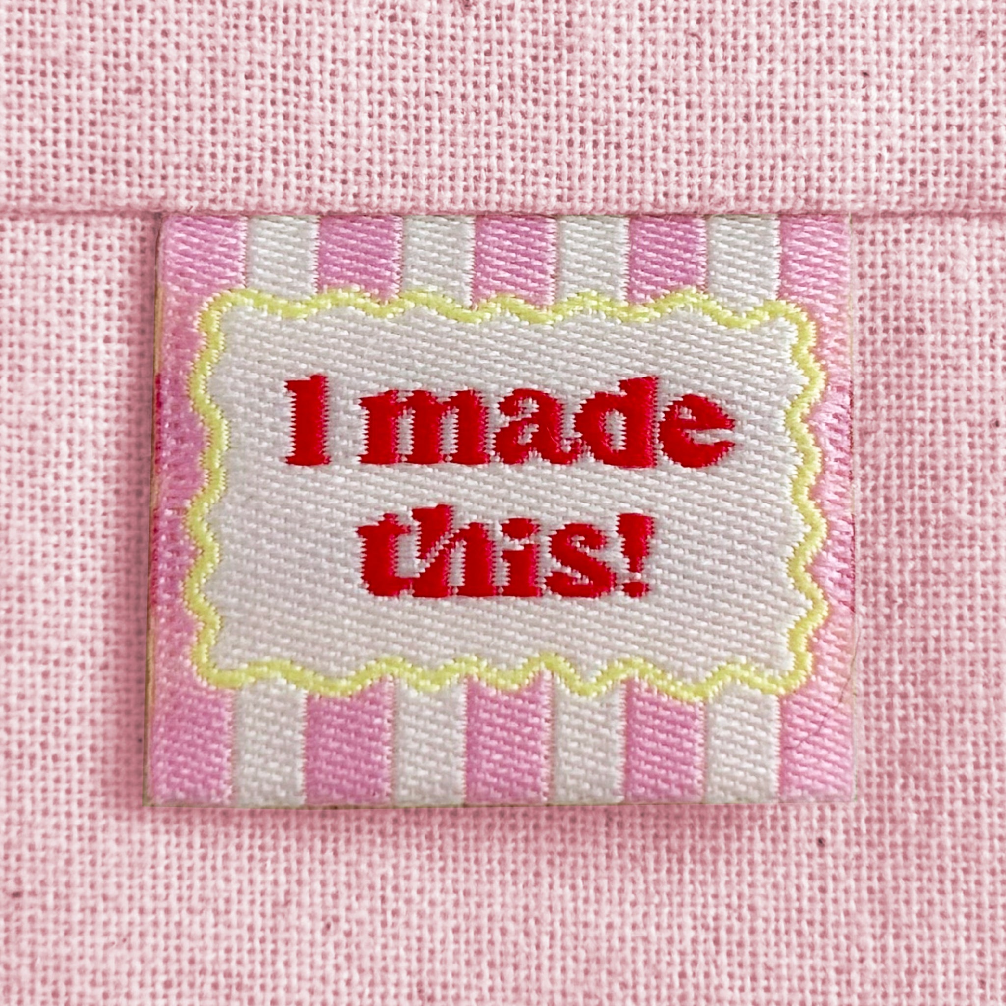 A close-up of a pink striped woven sewing label with a scalloped yellow border around red text reading 'I made this!'. The label is sewn into a light pink canvas fabric.