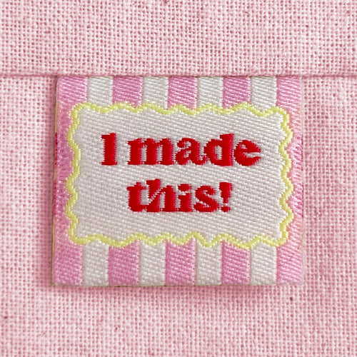 A close-up of a pink striped woven sewing label with a scalloped yellow border around red text reading 'I made this!'. The label is sewn into a light pink canvas fabric.
