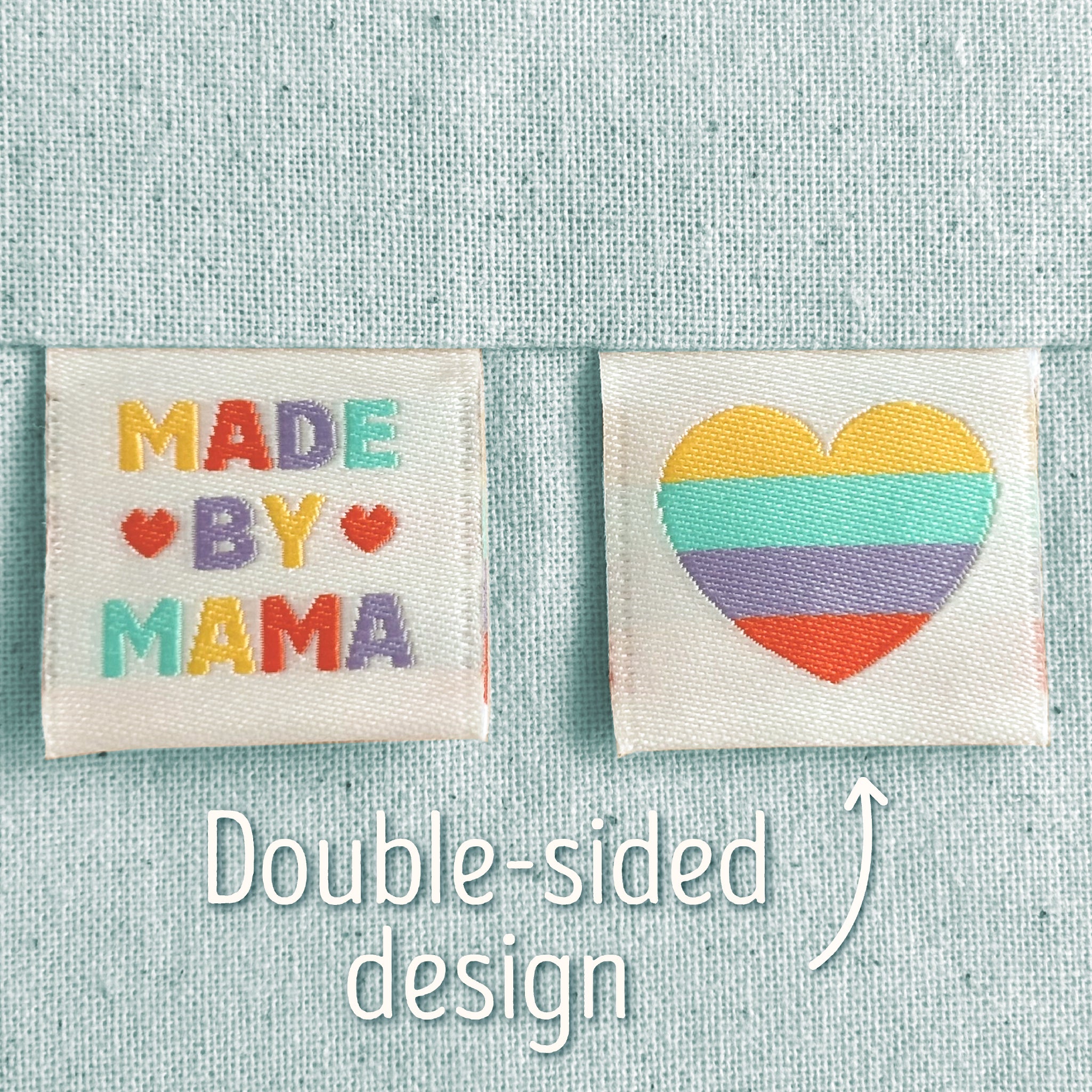 Two off-white, woven sewing labels that have been sewn into light blue canvas. The left patch reads “MADE BY MAMA” in multicoloured letters with small heart motifs, and the right patch shows a striped heart in yellow, teal, purple, and red. Text in the image explains that this is a double-sided design, implying that the heart is on the back of the 'Made By Mama' design.