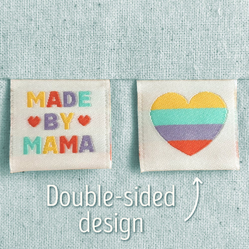 Two off-white, woven sewing labels that have been sewn into light blue canvas. The left patch reads “MADE BY MAMA” in multicoloured letters with small heart motifs, and the right patch shows a striped heart in yellow, teal, purple, and red. Text in the image explains that this is a double-sided design, implying that the heart is on the back of the 'Made By Mama' design.