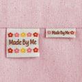 Two off-white, woven sewing labels sewn into light pink canvas fabric. The labels read "Made By Me" with small flower motifs in pink, red, and yellow.
