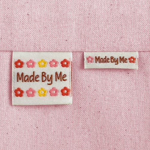 Two off-white, woven sewing labels sewn into light pink canvas fabric. The labels read "Made By Me" with small flower motifs in pink, red, and yellow.
