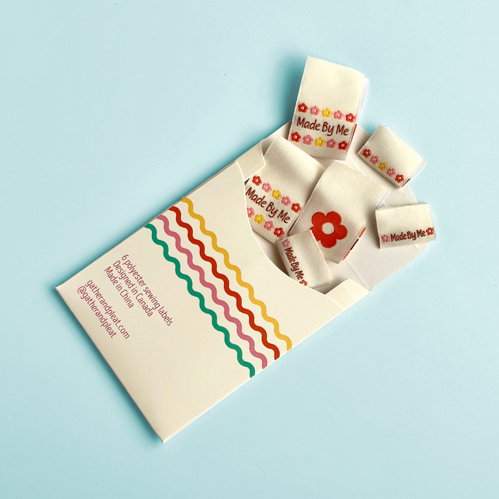 'Made by Me' woven sewing labels displayed beside opened Gather & Pleat packaging showing all six labels included in the pack.