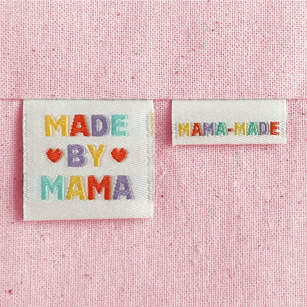 Two off-white embroidered woven sewing labels on a pink textured background. The larger label reads “MADE BY MAMA” with colourful embroidered letters and red hearts, and the smaller label reads “MAMA-MADE.