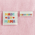 Two off-white embroidered woven sewing labels on a pink textured background. The larger label reads “MADE BY MAMA” with colourful embroidered letters and red hearts, and the smaller label reads “MAMA-MADE.