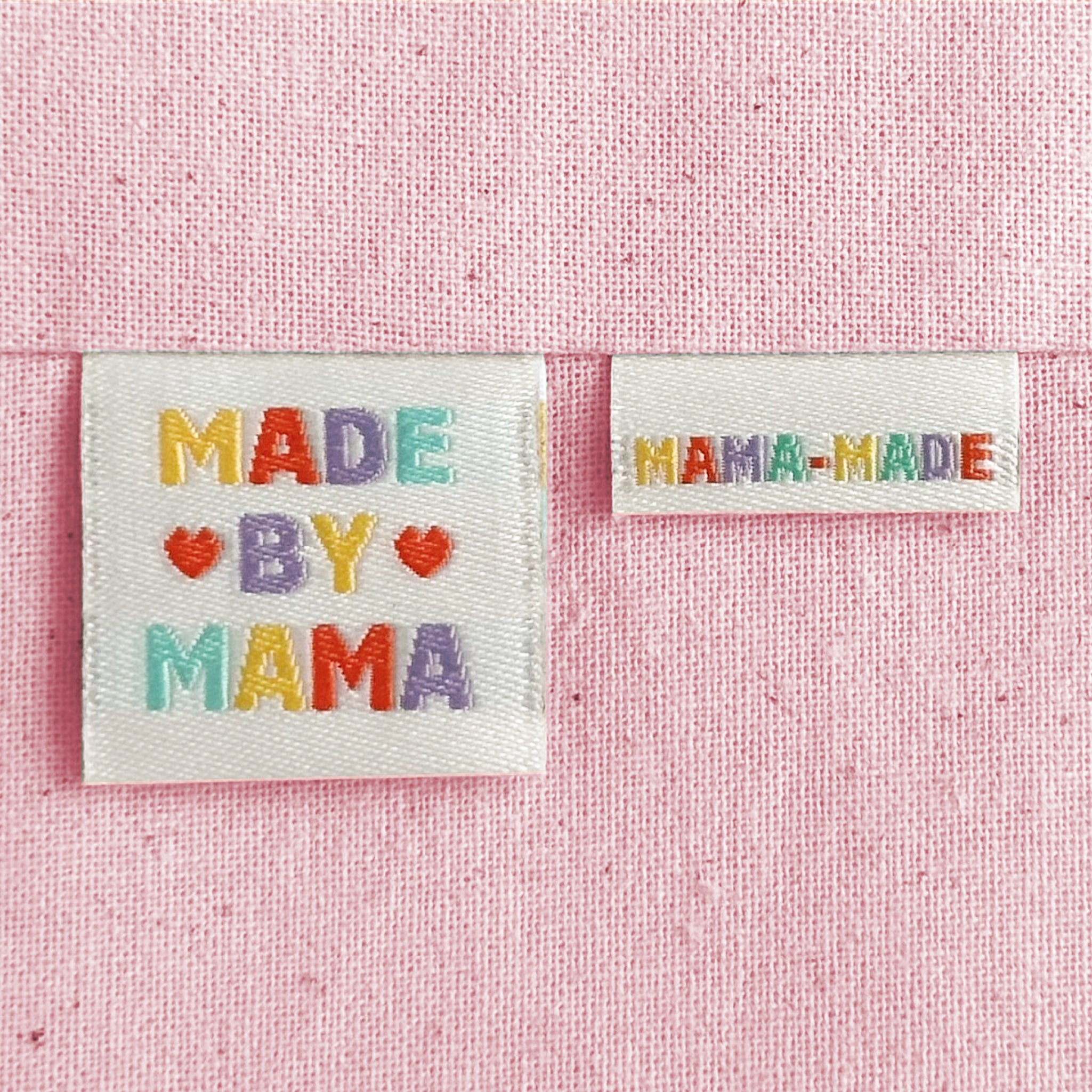 Two off-white embroidered woven sewing labels on a pink textured background. The larger label reads “MADE BY MAMA” with colourful embroidered letters and red hearts, and the smaller label reads “MAMA-MADE.