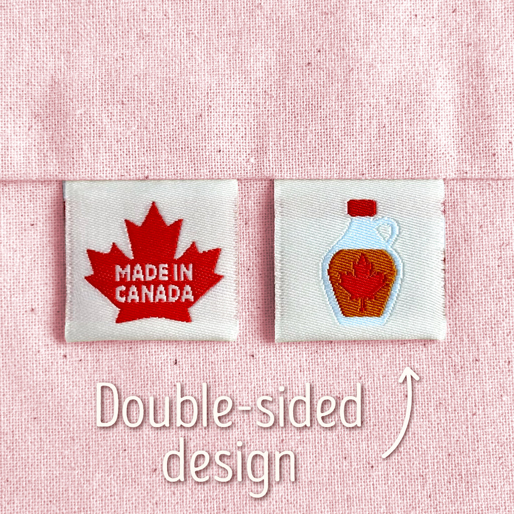 Two off-white, woven sewing labels that have been sewn into light pink canvas. The left patch reads “MADE IN CANADA” in white letters on a red maple leaf, and the right patch shows a bottle of maple syrup. Text in the image explains that this is a double-sided design, implying that the maple syrup bottole is on the back of the 'Made In Canada' design.