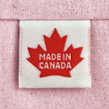 A close-up of a woven sewing label sewn into light pink woven fabric, featuring a red maple leaf with the words 'MADE IN CANADA'.