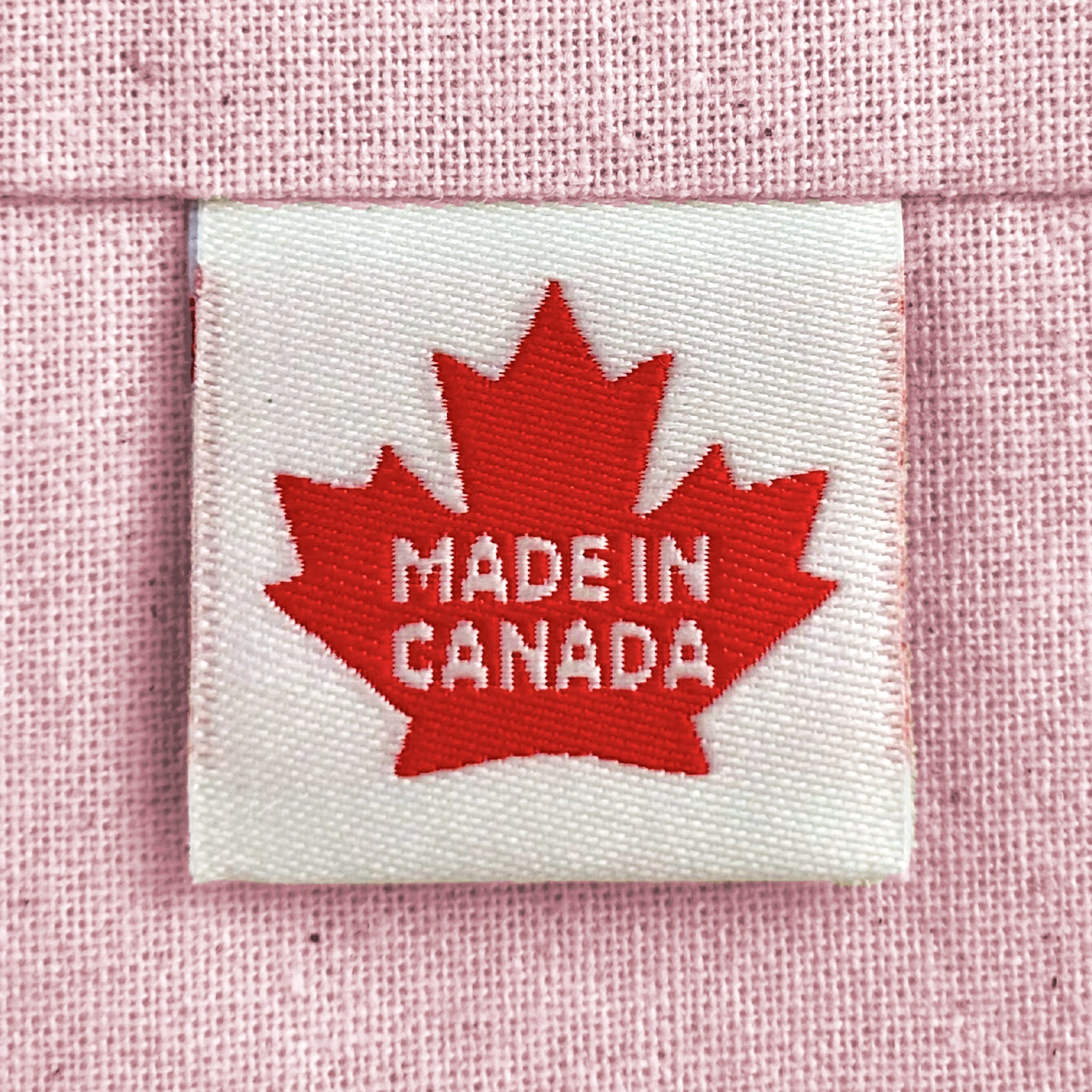 A close-up of a woven sewing label sewn into light pink woven fabric, featuring a red maple leaf with the words 'MADE IN CANADA'.