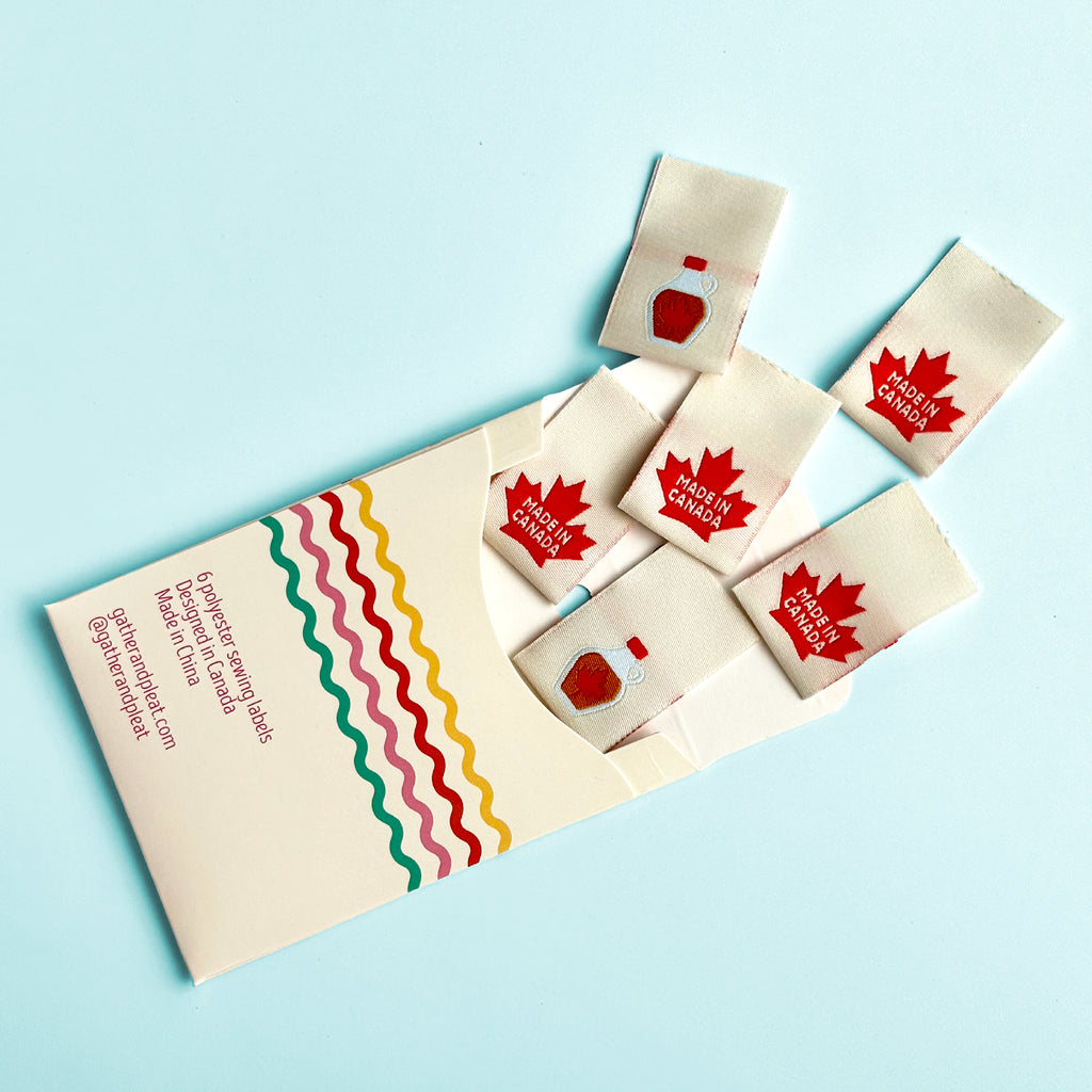 'Made in Canada' woven sewing labels displayed beside opened Gather & Pleat packaging showing all six labels included in the pack.