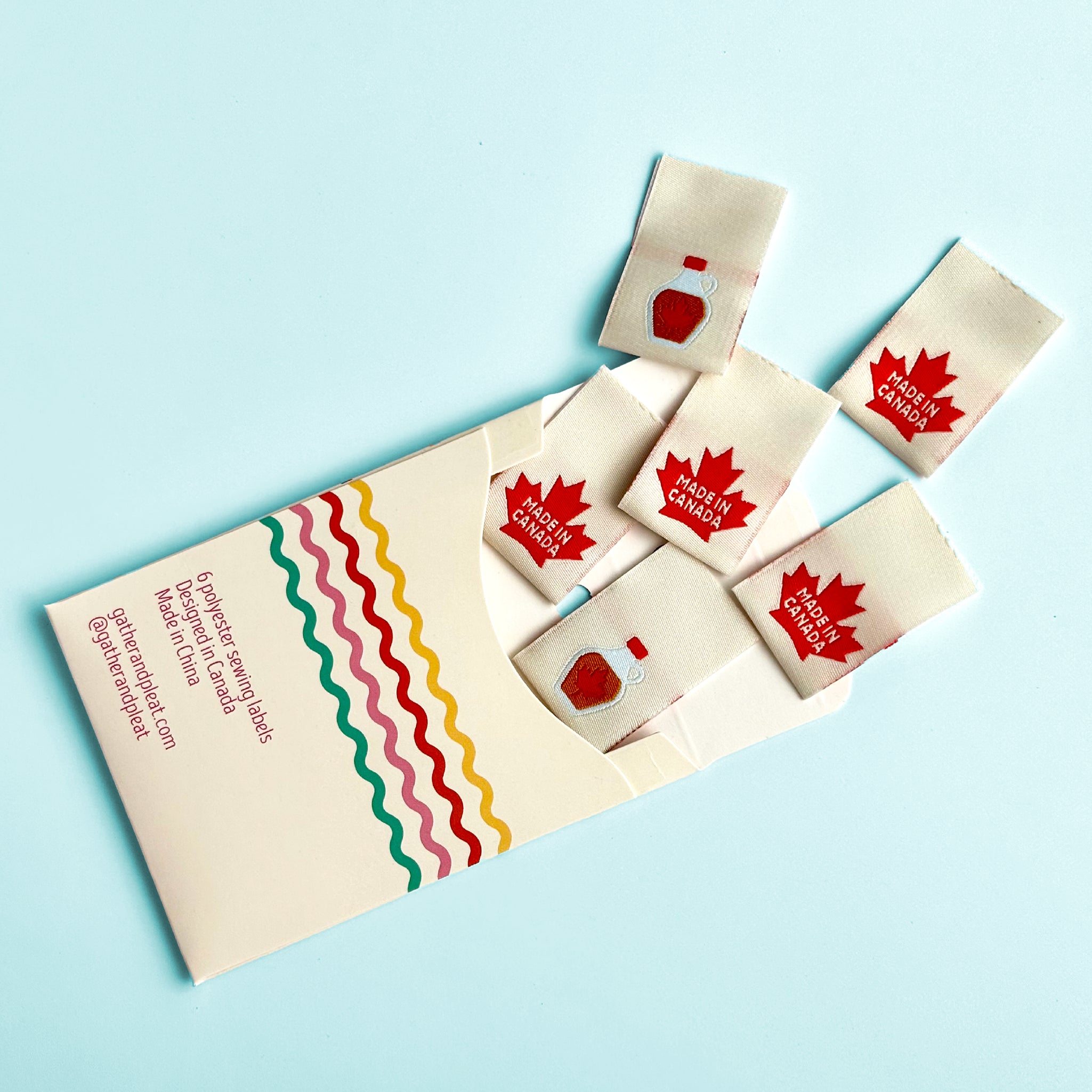 'Made in Canada' woven sewing labels displayed beside opened Gather & Pleat packaging showing all six labels included in the pack.