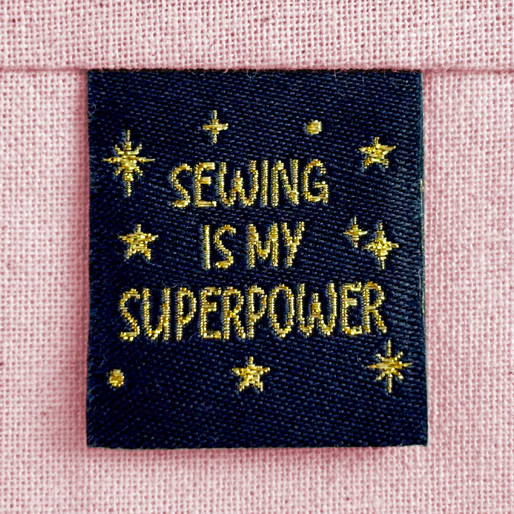 A black woven sewing label with sparkly gold lettering that reads "Sewing Is My Superpower". The label is sewn into light pink canvas.