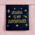 A black woven sewing label with sparkly gold lettering that reads "Sewing Is My Superpower". The label is sewn into light pink canvas.