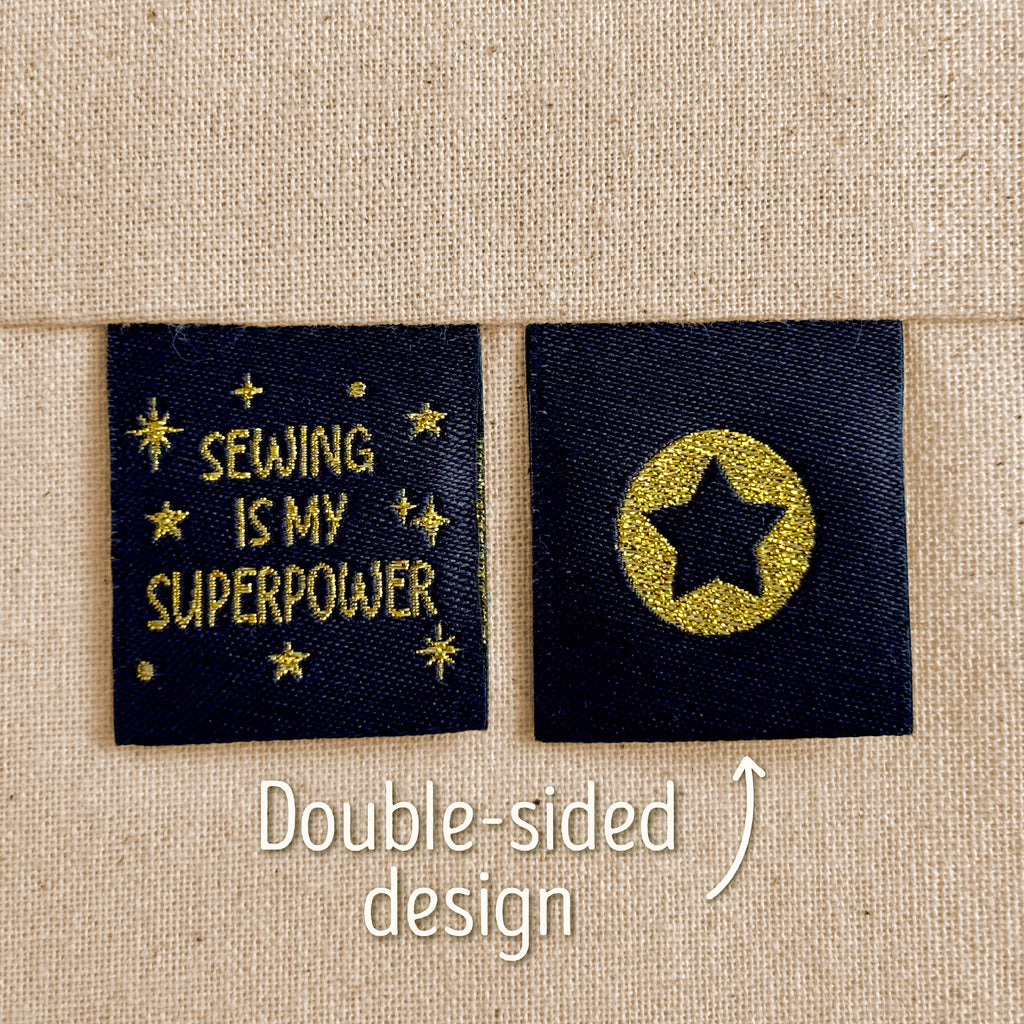 Two black woven sewing labels that have been sewn into beige canvas. The left label reads “Sewing Is My Superpower” in sparkly gold lettering, which surround by gold stars. The right label shows a black star inside a sparkly gold circle. Text in the image explains that this is a double-sided design, implying that the star is on the back of the 'Sewing Is My Superpower' design.