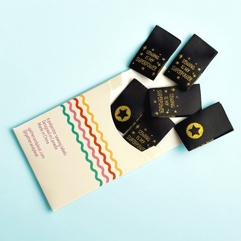 'Sewing Is My Superpower' woven sewing labels displayed beside opened Gather & Pleat packaging showing all six labels included in the pack.
